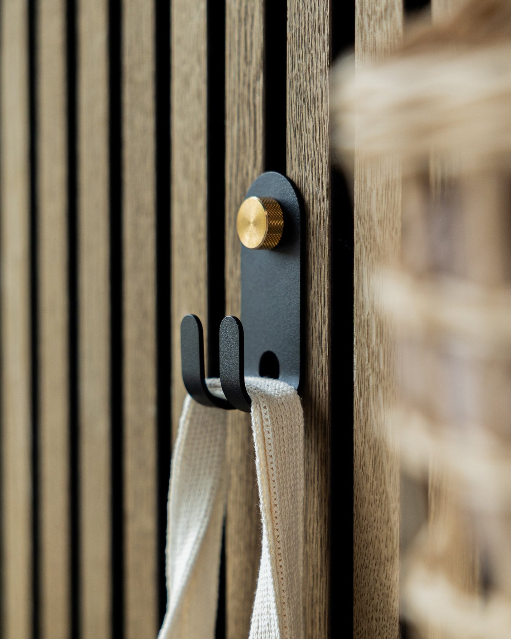 De WoodUpp Norr Double Hanger in black is gemonteerd op een akoestisch lattenwandpaneel van Nordic hout en houdt een crèmekleurige stoffen lus vast.