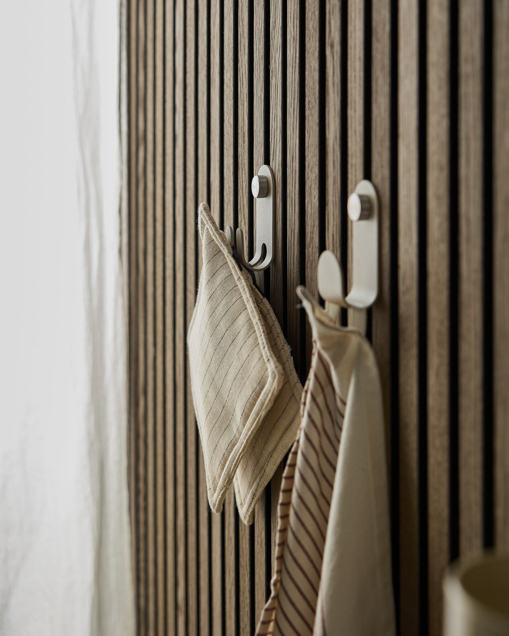 Twee gestreepte keukendoeken hangen aan de Norr Double Hanger van WoodUpp, gemonteerd op een beige Scandinavische lattenwand Akoestisch paneel, verlicht door zacht natuurlijk licht.