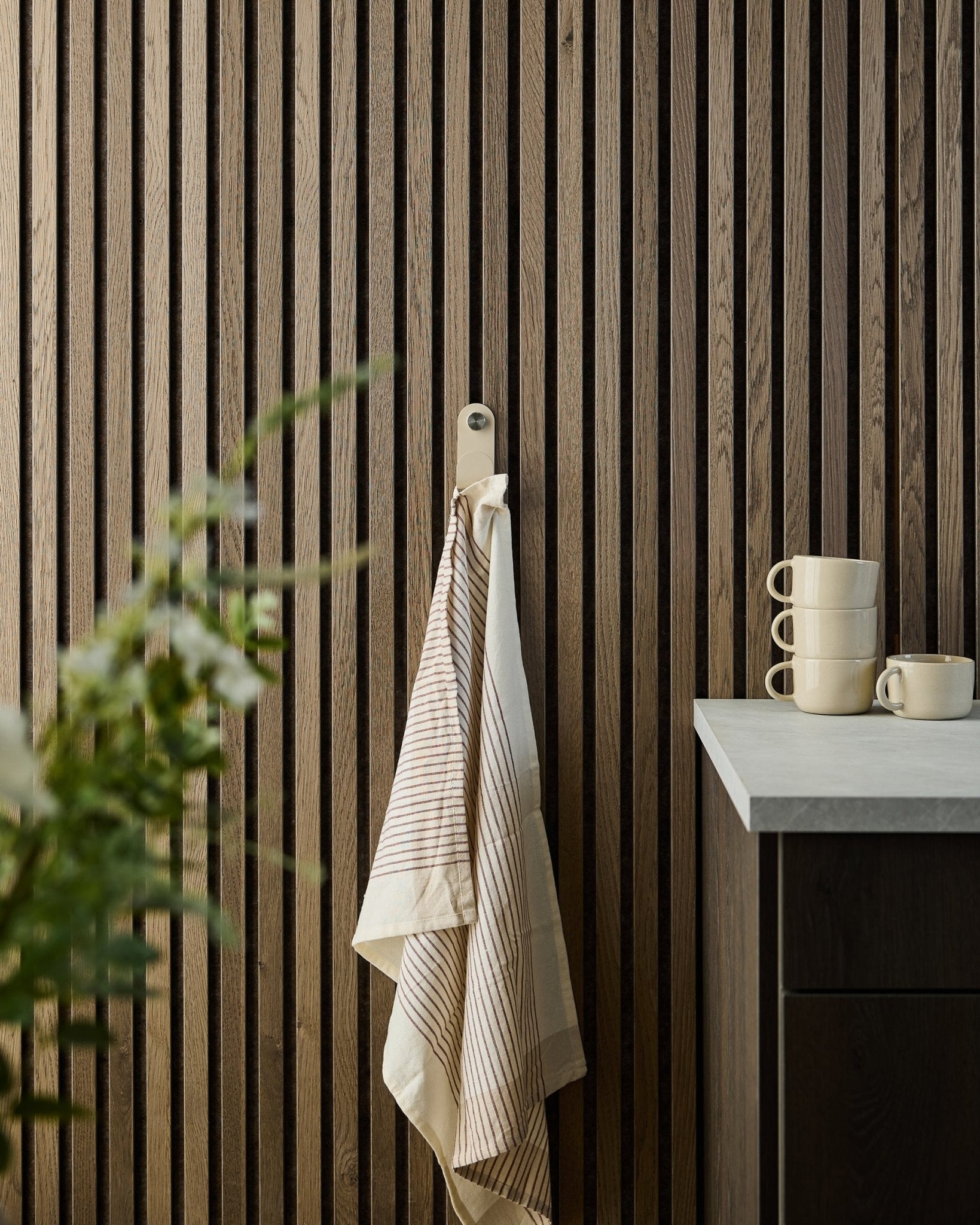 Een gestreepte handdoek hangt aan een beige WoodUpp Norr Single Hanger, gemonteerd op een houten lattenpaneel Akoestisch paneel; op het aanrecht eronder staan gestapelde mokken en een plant.