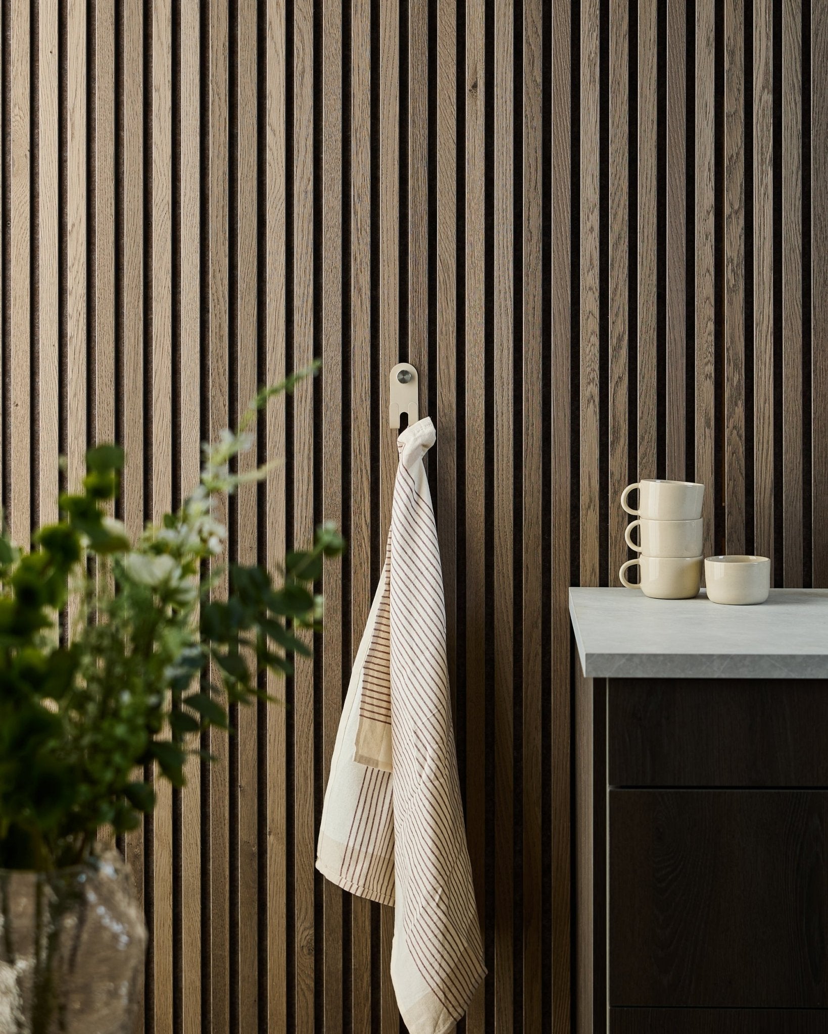 Een gestreepte handdoek hangt aan de WoodUpp Norr Double Hanger in beige, gemonteerd op een Scandinavisch akoestisch lattenwandpaneel; mokken, een kom en een groene plant staan eronder.