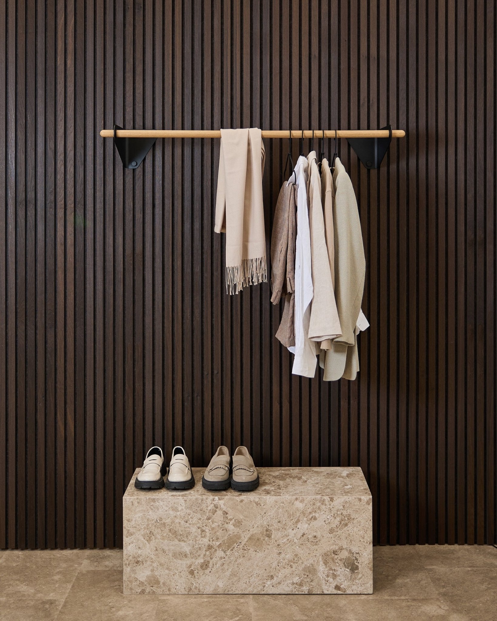 Kleding in neutrale tinten en een sjaal hangen aan een black van 115,9 cm Linn | Coat Rail van WoodUpp boven een marmeren bank, tegen een donker Akupanel akoestisch lattenwandpaneel.