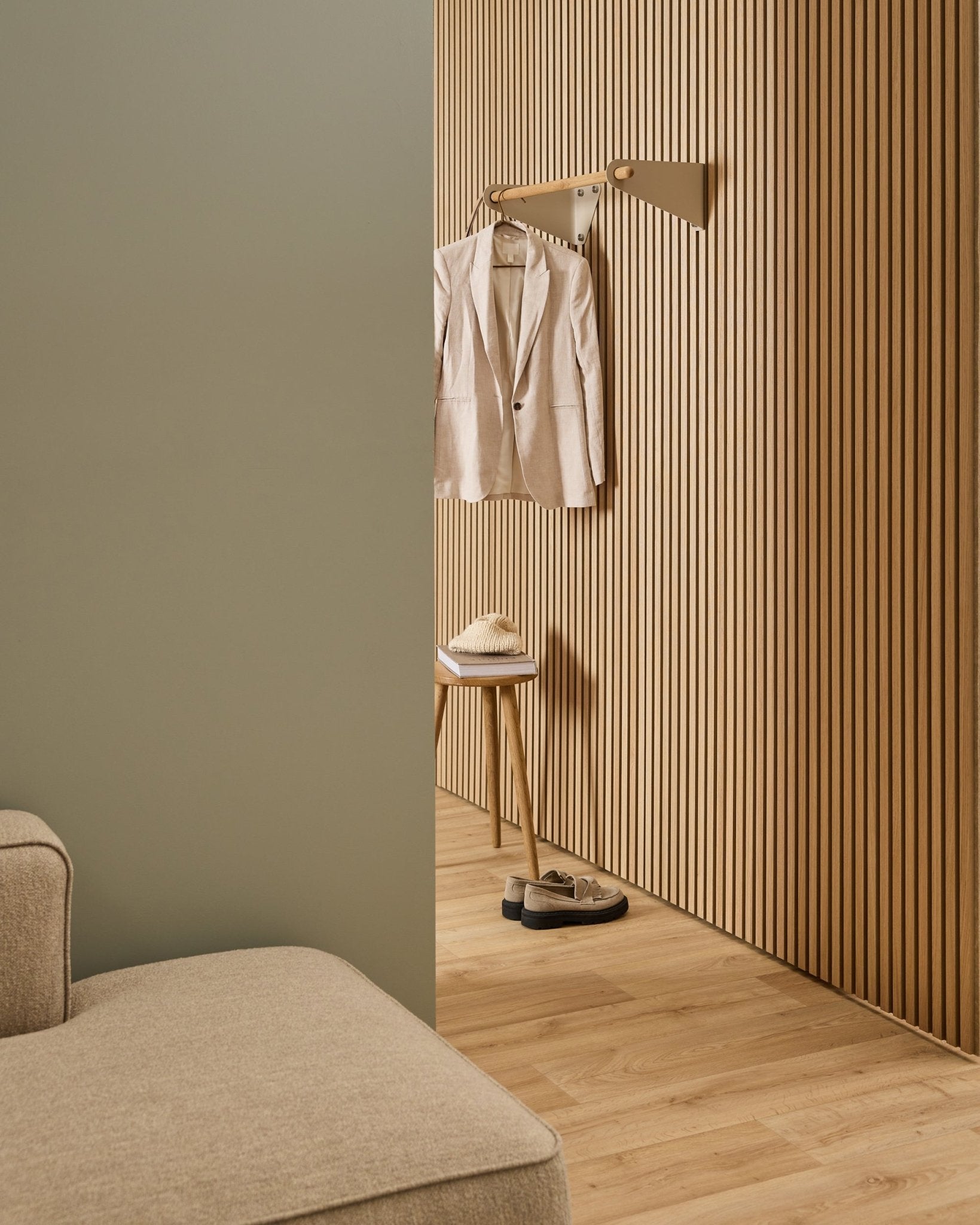 Minimalistische ruimte met beige bank, Akupanel akoestische lattenwand, WoodUpp Linn kapstokrail in beige (75,9 cm), kruk met hoed en schoenen op een licht houten vloer.