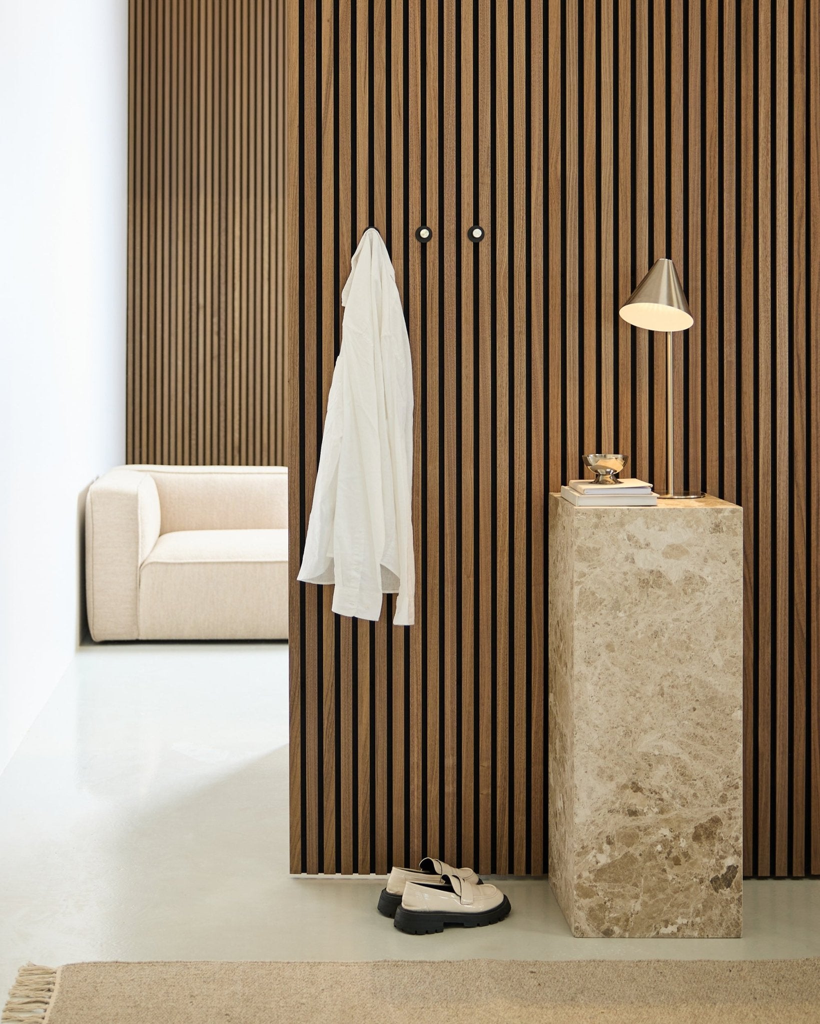 Minimalistische ruimte met een Scandinavische akoestische wand van houten latten in Akupanel, een wit overhemd aan wandhaken, een stenen sokkel met lamp en boeken, een beige bank en witte schoenen op de vloer.