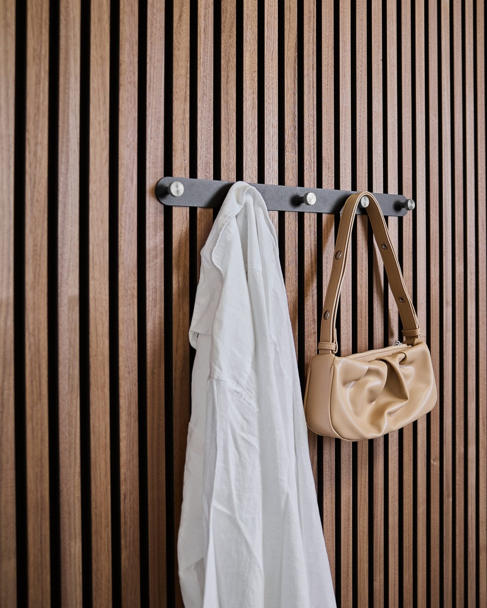 Une chemise blanche et un sac à main beige sont suspendus aux crochets muraux Black/Stainless Steel X5 Norr de WoodUpp, fixés sur un panneau mural acoustique à lattes vertical.