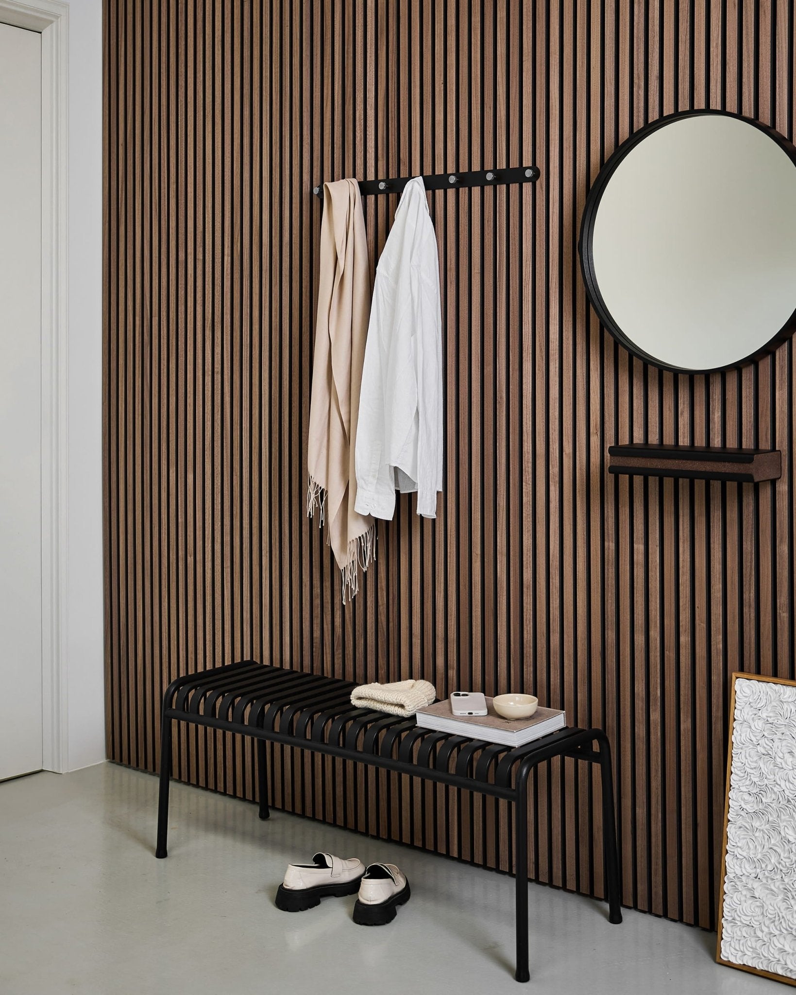 Moderne entree met een Scandinavische wand van akoestische houten lattenpanelen, ronde spiegel, black bankje, WoodUpp Norr kapstok en pantoffels op een lichte vloer.