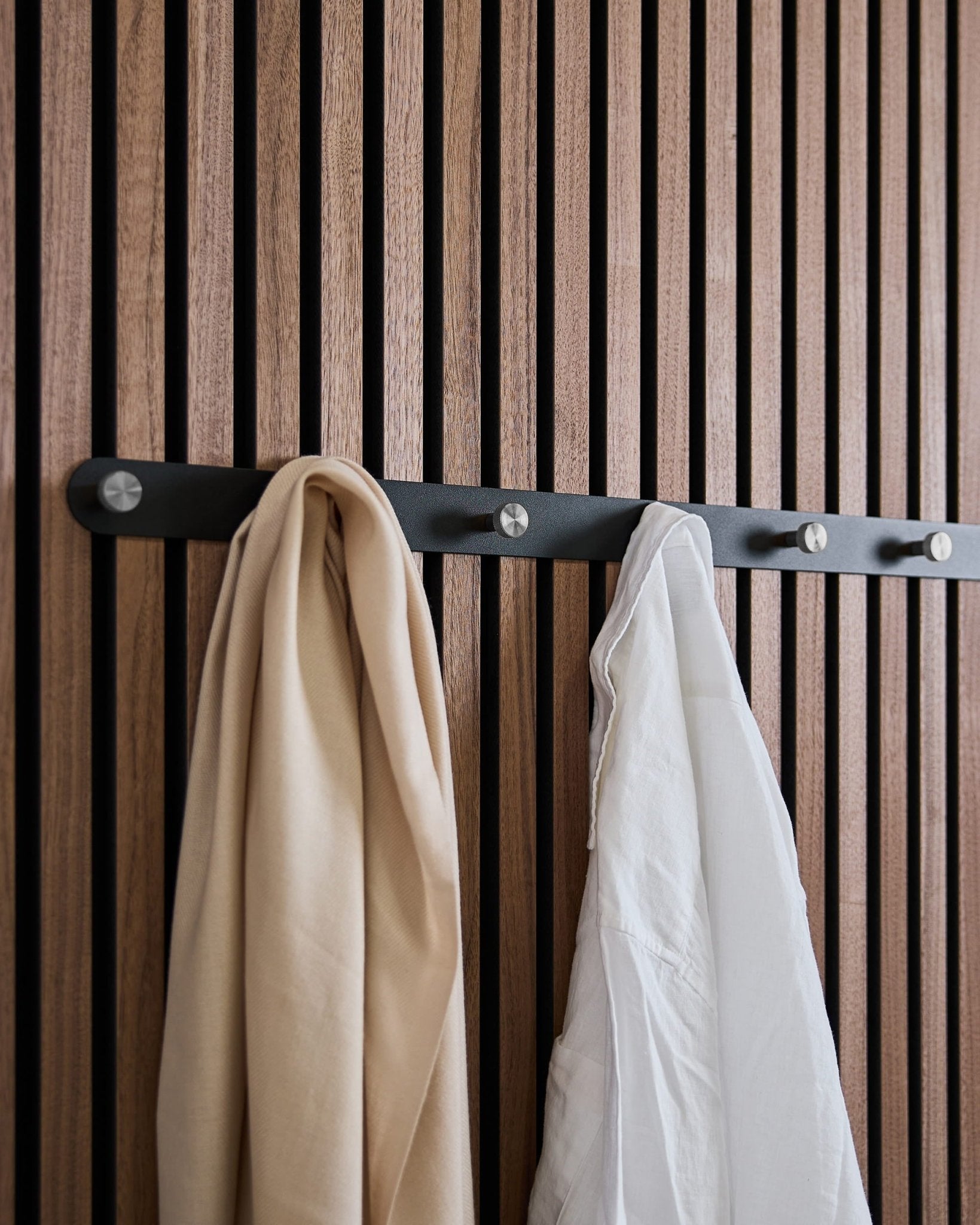 Een beige sjaal en wit overhemd hangen aan WoodUpp’s Norr X7 Black/Stainless Steel kapstok, gemonteerd op een Scandinavisch akoestisch lattenwandpaneel.