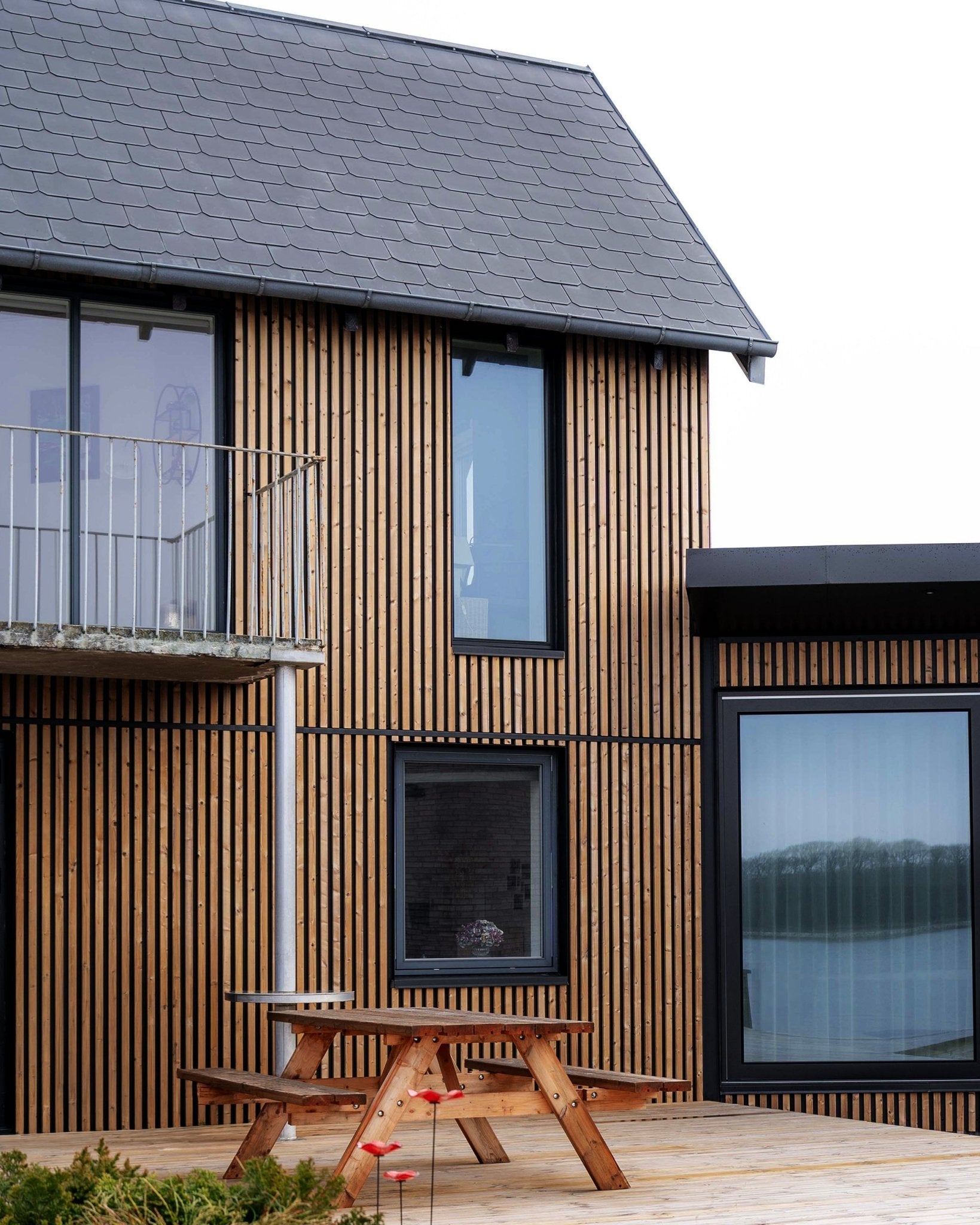 Moderne woning met WoodUpp Aluwood gevelpanelen in geolied Brown, met large ramen, een balkon en een terras met een houten picknicktafel.