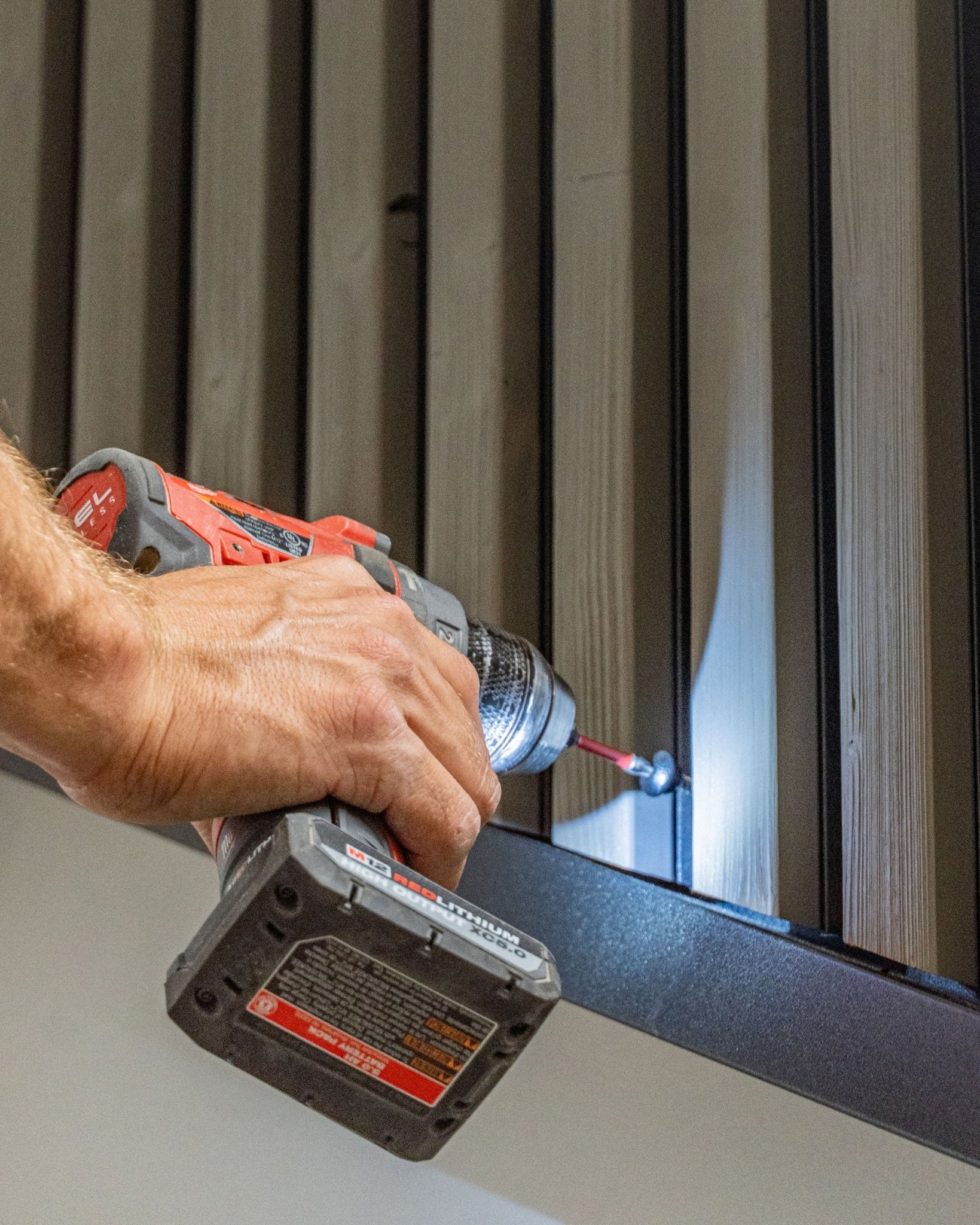 Une personne fixe une vis de façade black WoodUpp Front Screw (4,8x35, rondelle Ø9,5) dans un panneau de façade vertical Aluwood à l’aide d’une perceuse-visseuse sans fil.