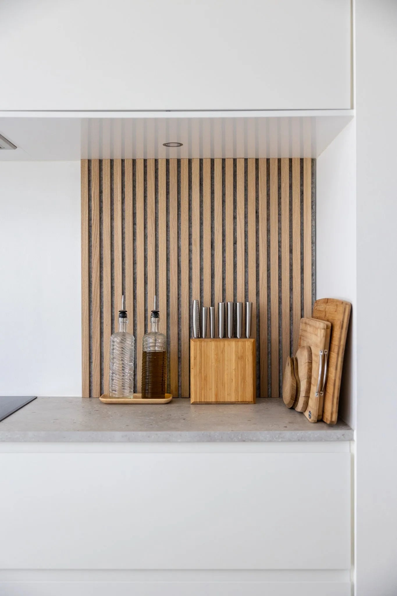 Comptoir de cuisine moderne avec bouteilles dhuile, bloc de couteaux en bois et planches à découper empilées sur un dosseret en lattes de bois acoustique Akupanel nordique et des armoires blanches.