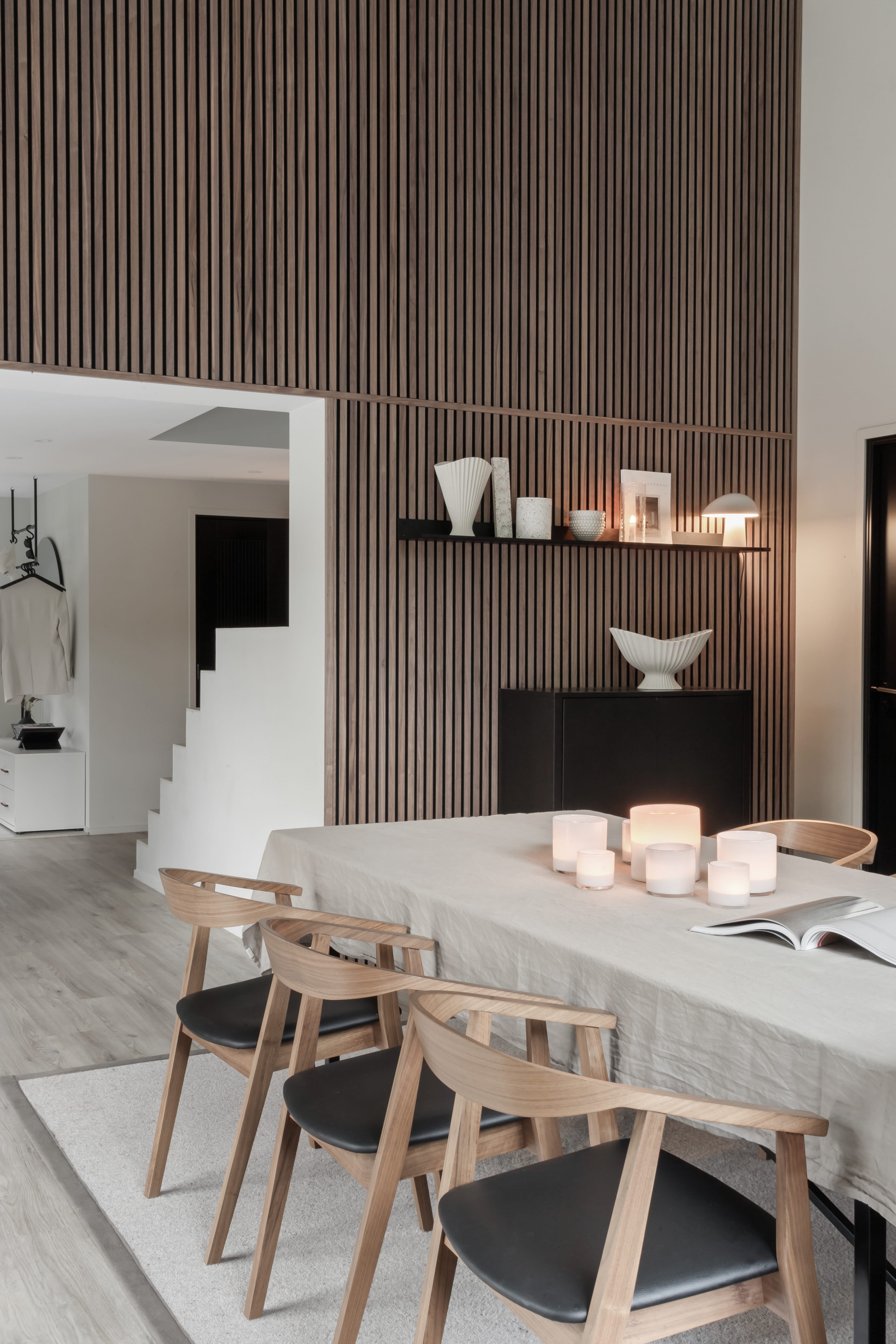 Salle à manger moderne avec une table en bois clair, quatre chaises, des bougies et un décor minimaliste. Mur vertical en lattes de bois acoustique Akupanel et escalier ouvert en vue.
