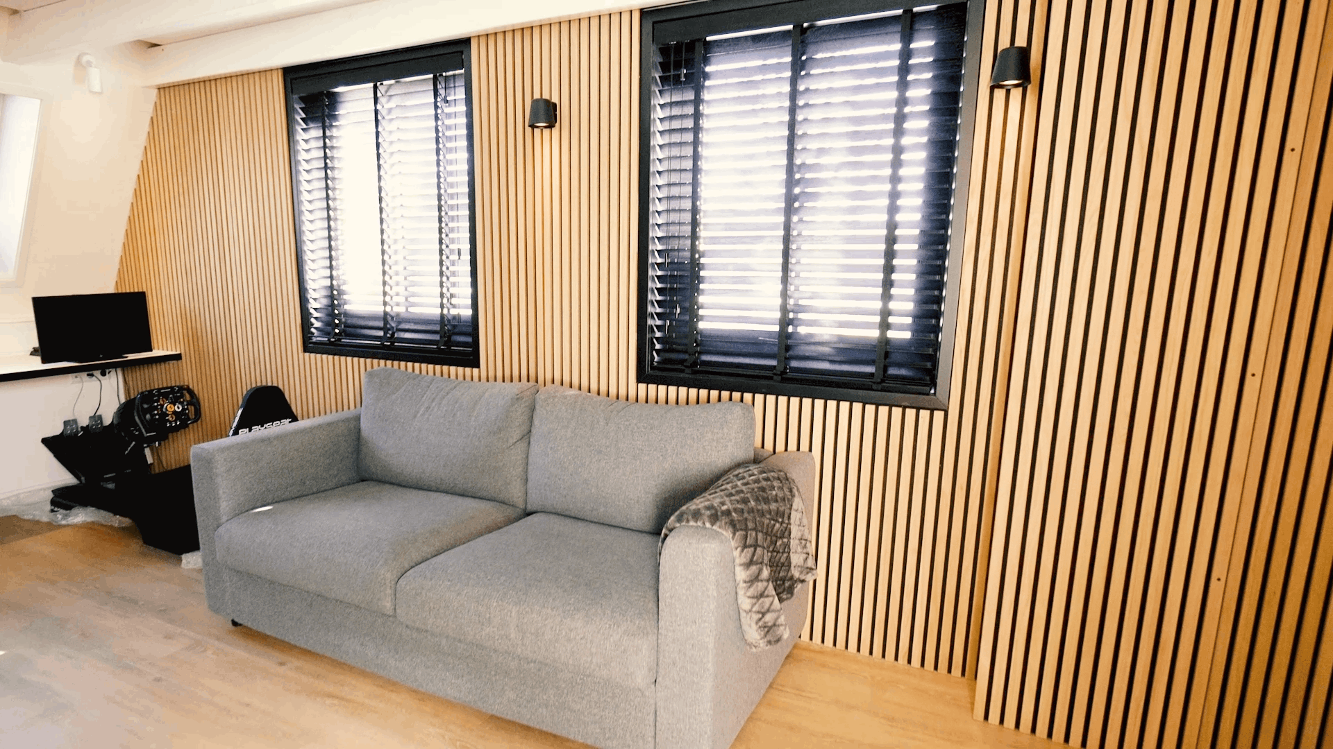 Canapé gris avec couverture à motifs placé devant un mur de lattes en bois acoustique Akupanel nordique, flanqué de deux fenêtres avec stores noirs et dun bureau compact.