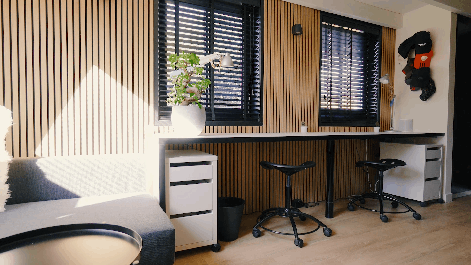 Bureau moderne avec des panneaux acoustiques en bois nordique sur les murs, des postes de travail ergonomiques, des tabourets noirs, une plante en pot et la lumière du soleil à travers les stores noirs.