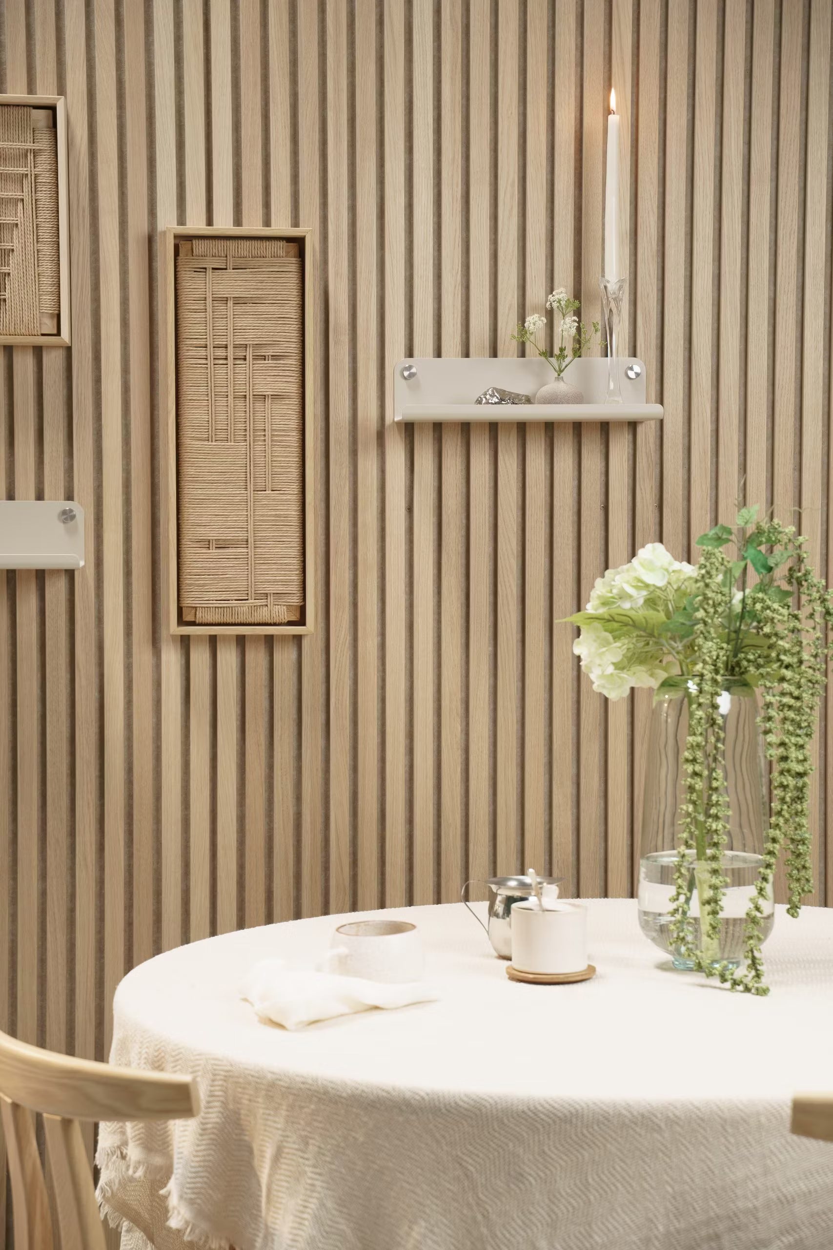 Een ronde tafel met een wit kleed, bloemenvaas, kaars en decor geplaatst voor een lichthouten Akupanel akoestische lamellenwand met geweven en plankenaccenten.