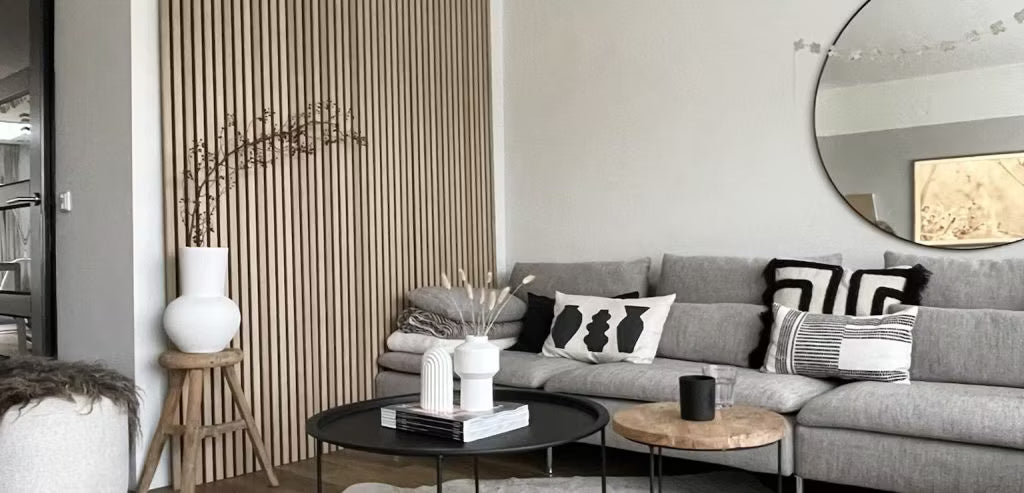 Moderne woonkamer met een grijze bank, abstracte kussens, zwarte salontafels, decoratieve vazen en een akoestische Akupanel houten lamellenwand met een ronde spiegel.