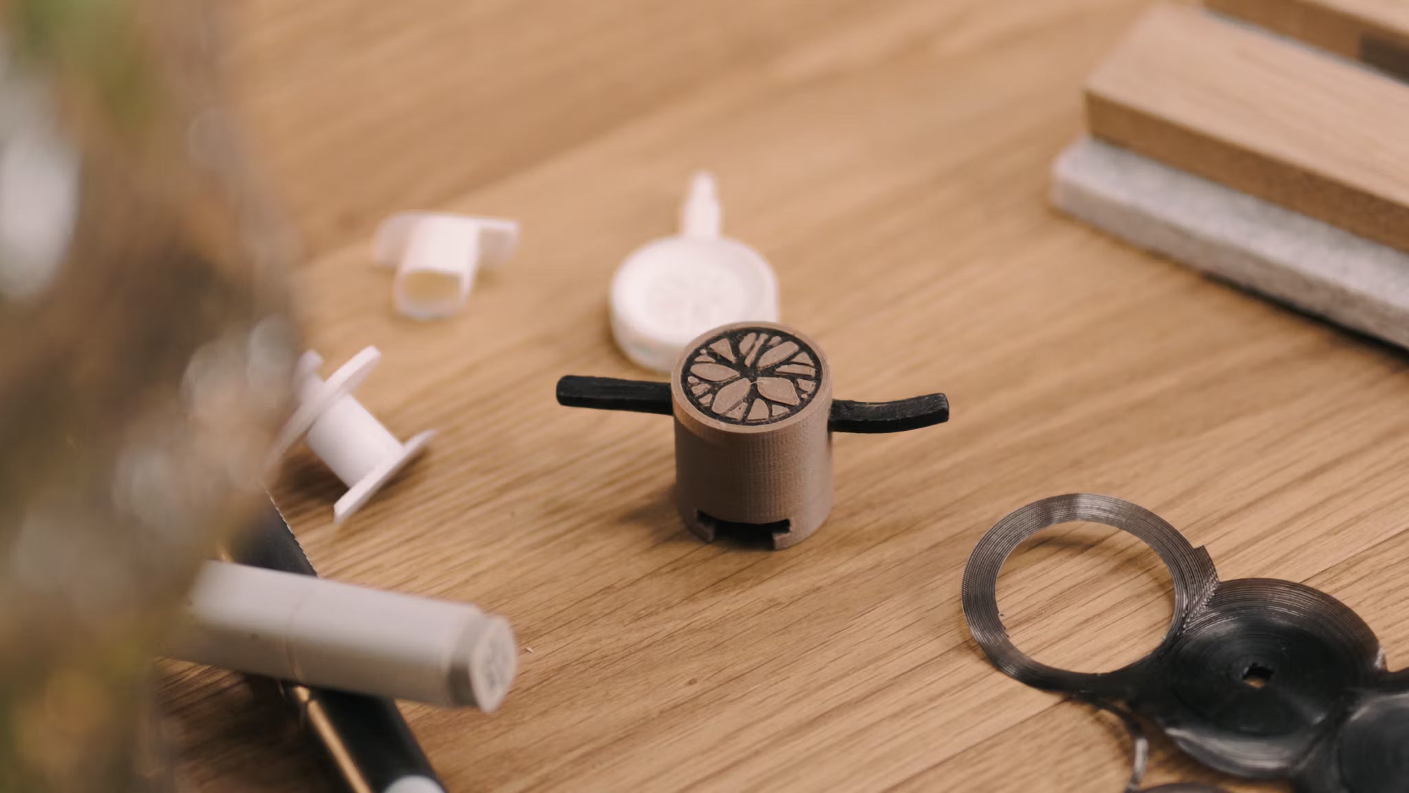 Een klein 3D-geprint prototype met zwarte armen staat op een houten bureau naast verschillende plastic onderdelen, verwijzend naar de ontwikkeling van accessoires voor akoestische panelen.