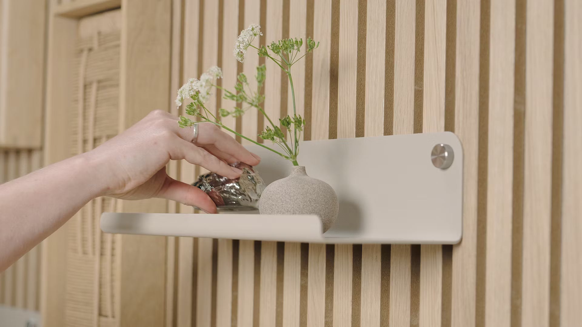 Een hand plaatst een klein decoratief object naast een vaas met witte bloemen op een minimalistische plank tegen een gestructureerd Scandinavisch akoestisch houten lattenpaneel.