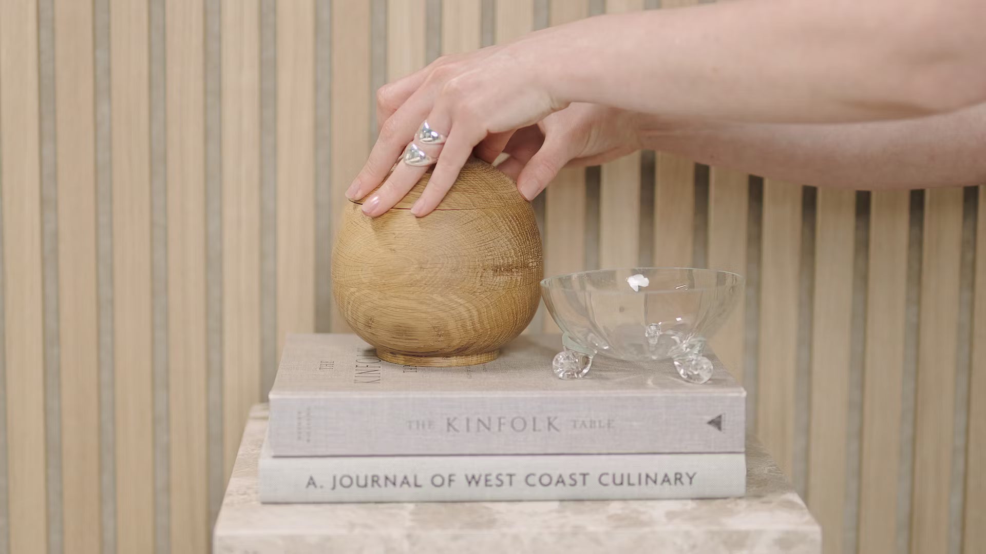 Een hand raakt een houten bol aan die op gestapelde boeken ligt, waaronder Kinfolk Table en Journal of West Coast Culinary, naast een glazen kom op een akoestisch paneel van houten latten.