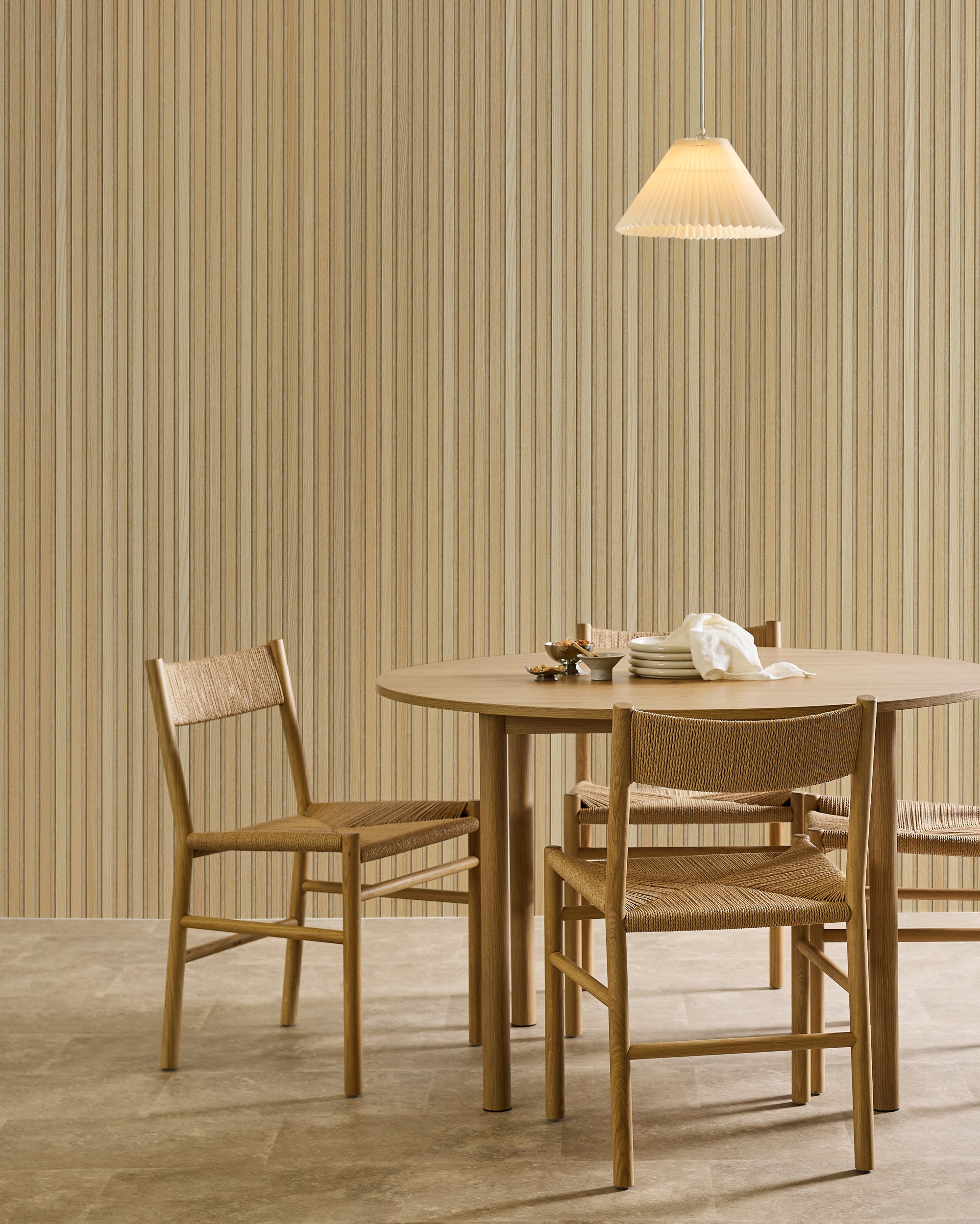 A round wooden dining table with three woven chairs, set with plates and cups, under a pendant light. Akupanel acoustic wood panels on the walls in a minimalist interior.