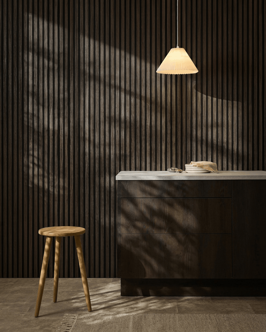 Minimalistische keuken met WoodUpp Akupanel Black Oak akoestische panelen aan de muur, een houten kruk, wit werkblad, beige hanglamp en schaduwen in het zonlicht.