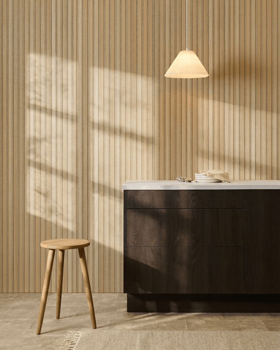 Scandinavische keuken met WoodUpp Akupanel Classic Oak akoestisch lattenwandpaneel, donkere houten kasten, houten kruk, hanglamp en zonlicht dat schaduwen werpt.
