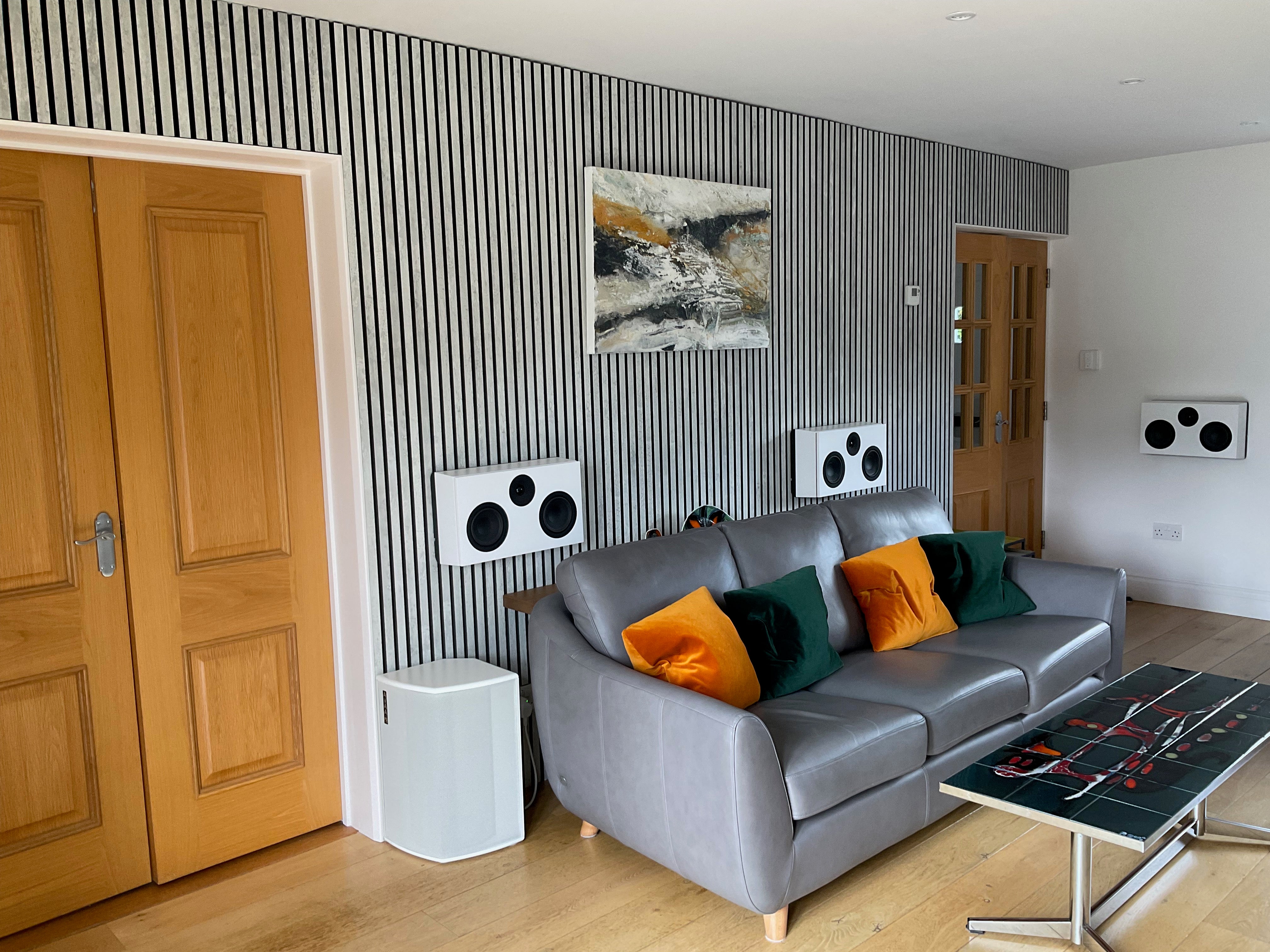 Modern living room with gray sofa, orange and green pillows, striped Akupanel accent wall, abstract art, and wall speakers for surround sound.