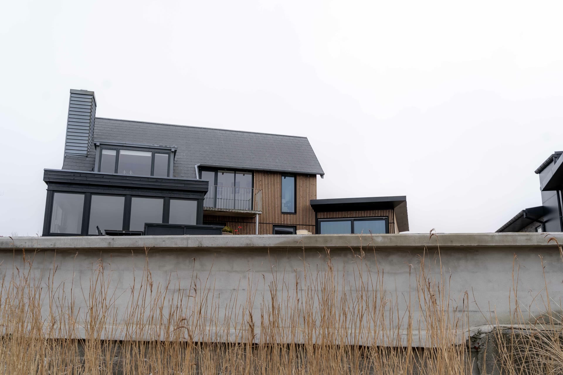 House with Aluwood slat cladding, dark slate roof and black glazed extension seen over a concrete sea wall and reeds
