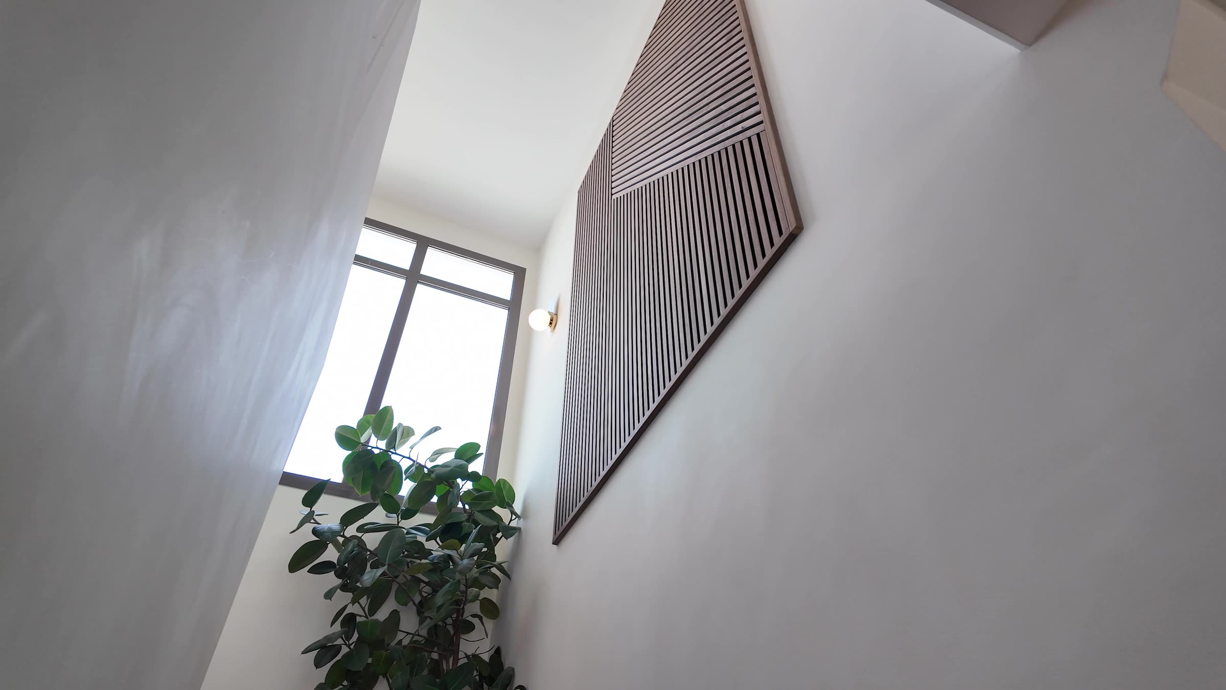 Looking up a stairwell at a large framed Akupanel slat artwork on the wall beside a tall window, a wall light and a plant
