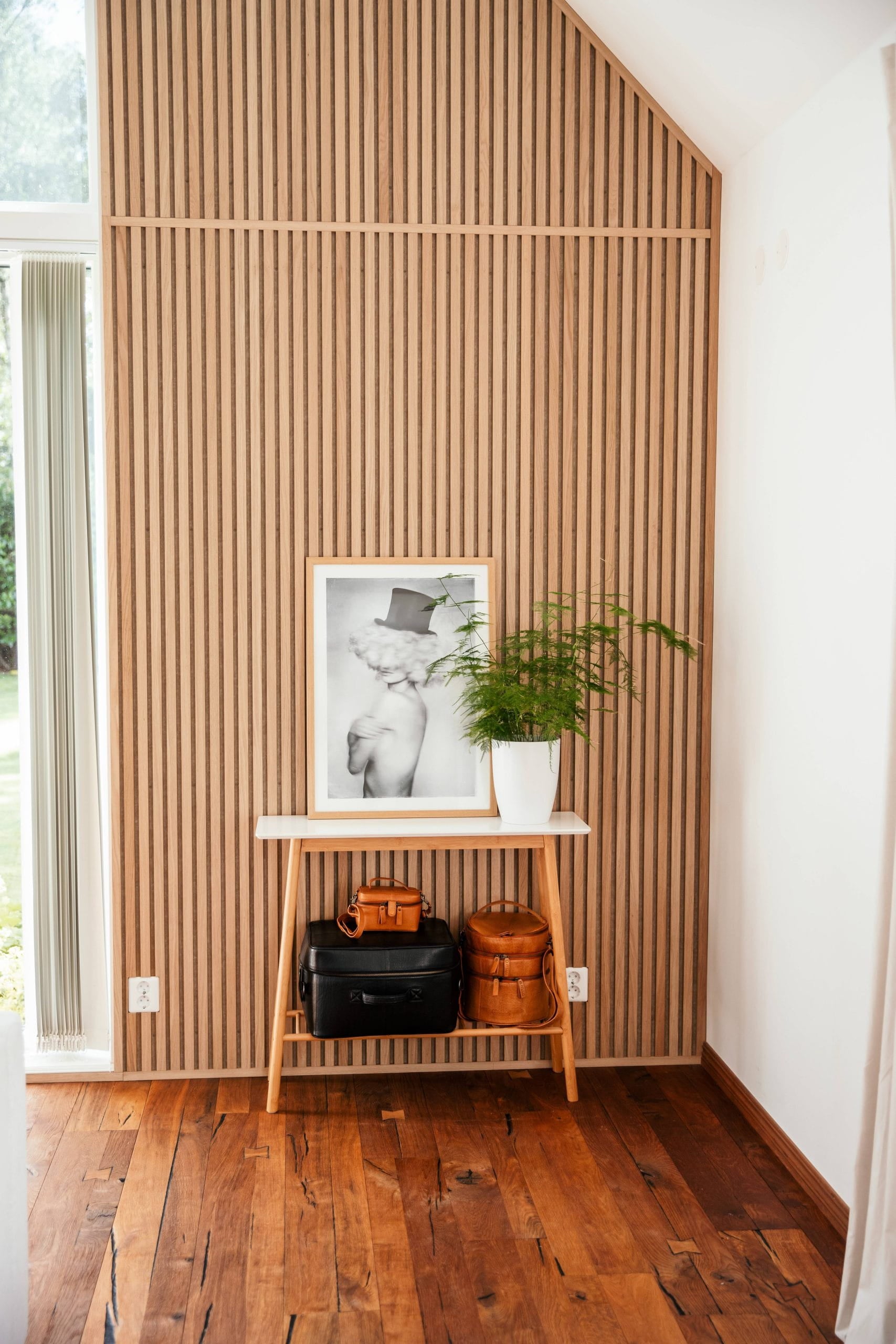 Een kleine houten tafel met tassen, een ingelijste foto en een potplant staat tegen een Scandinavisch akoestisch lamellenpaneel in een lichte kamer met houten vloeren.