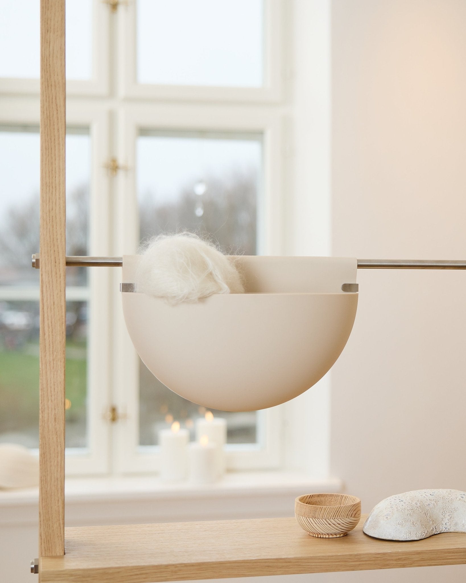 Een witte schaal met een pluizig object staat op een houten plank naast kaarsen, decoratie en een akoestisch accessoire Gro Soft Pocket BM2 van WoodUpp in licht beige bij het raam.