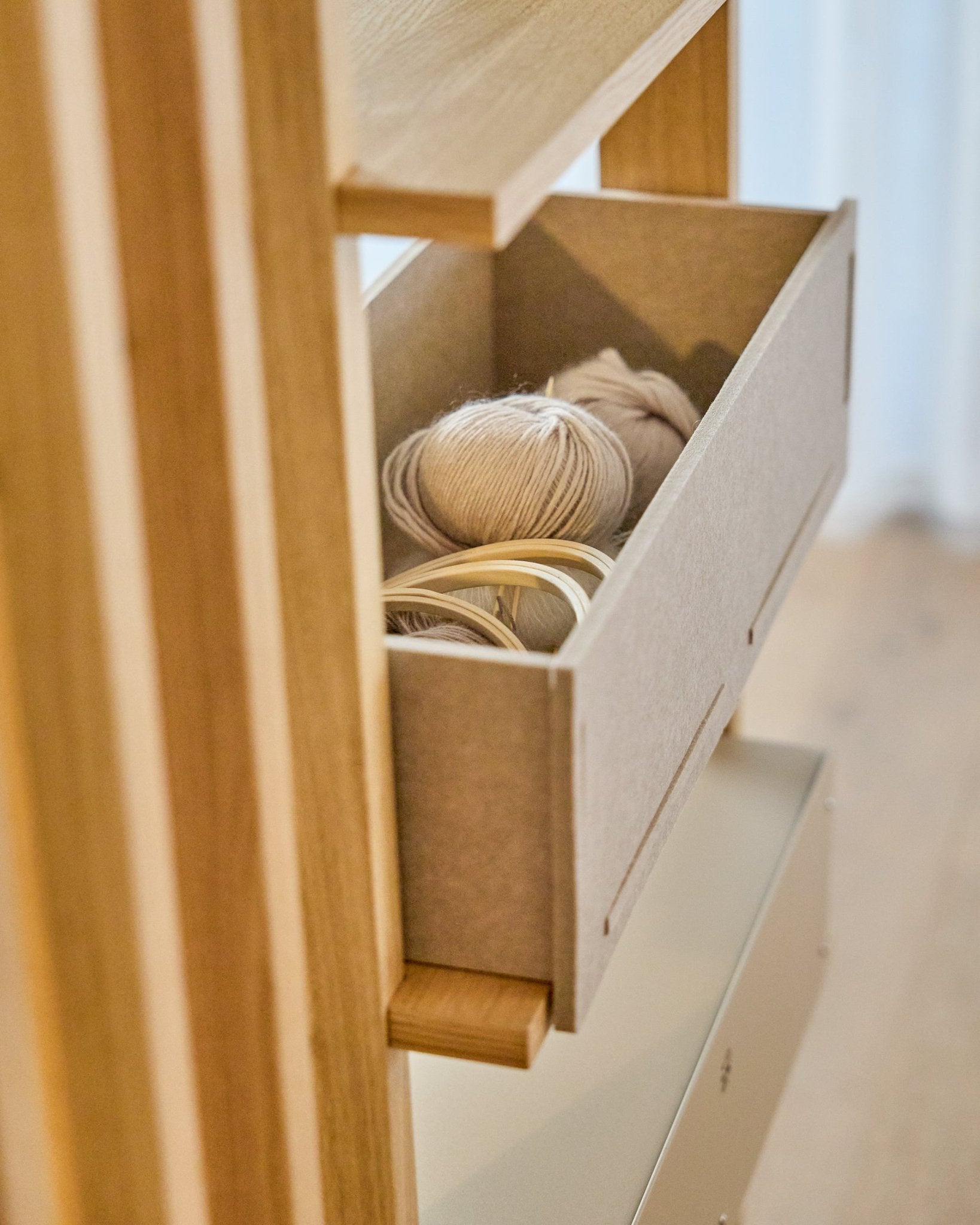 De WoodUpp Vigg Felt Box / BM2 in beige biedt plek aan lichte bollen garen en houten borduurringen op een houten wandrek, als aanvulling op Scandinavische akoestische panelen.