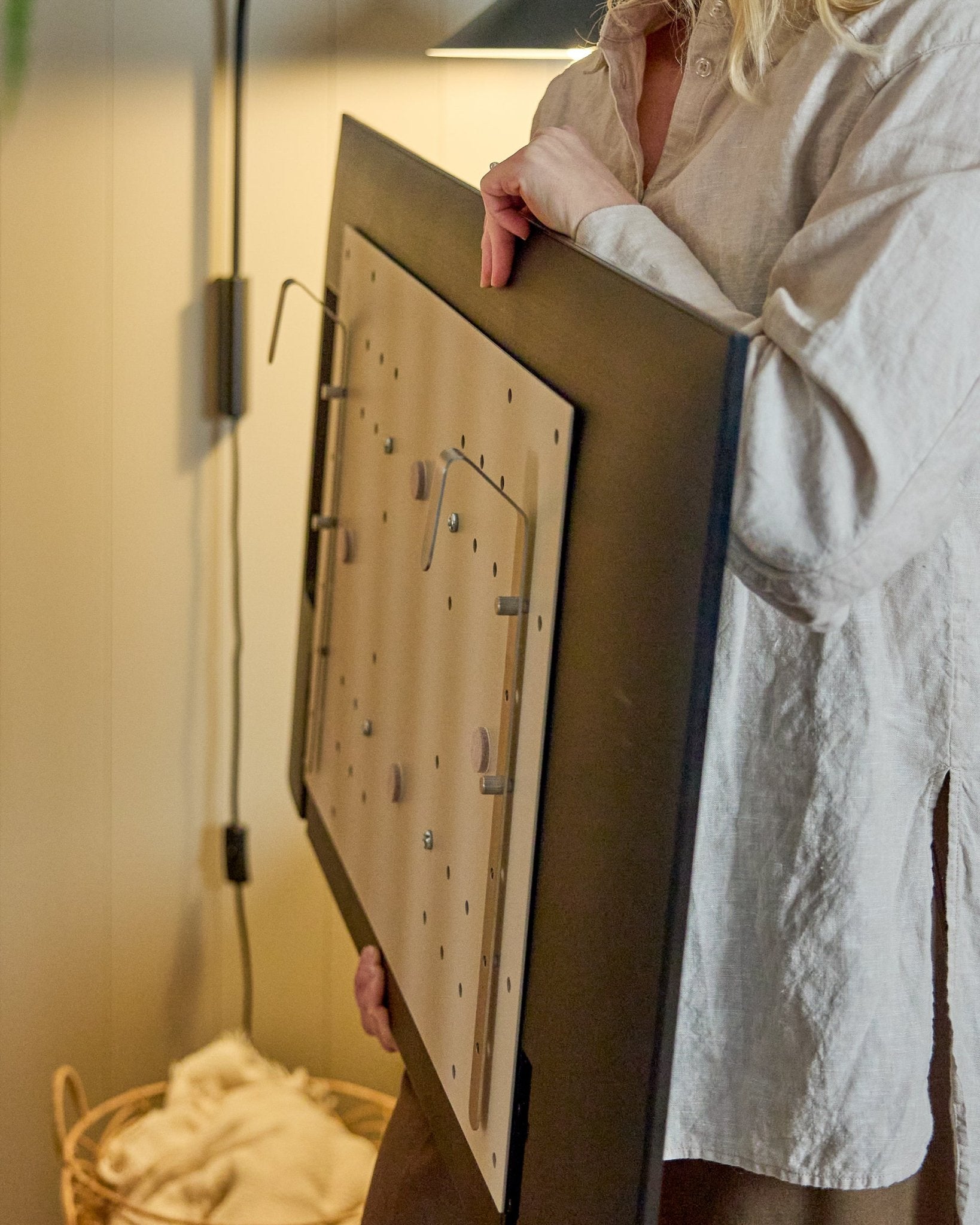 Une personne en chemise claire tient le support TV WoodUpp Nell BM1 en beige, 64x44 cm, compatible avec les panneaux acoustiques en bois nordiques, pour des installations sans mur.