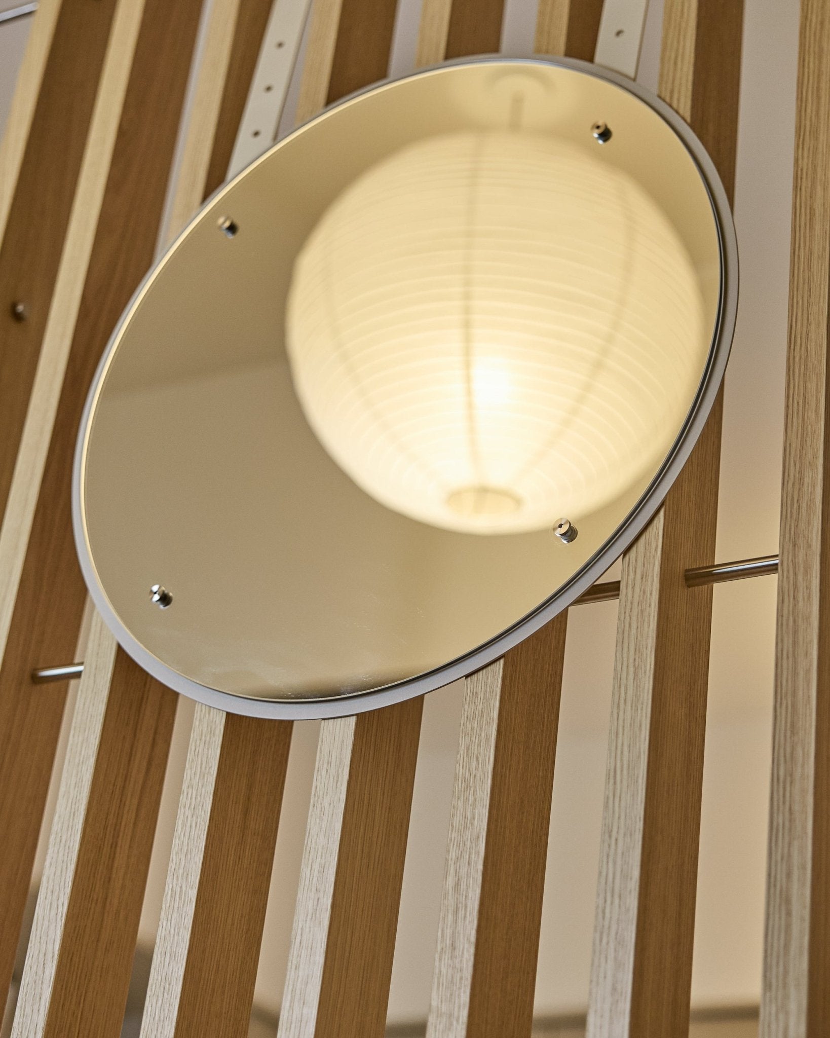 De Vala | Mirror / BM1, D54 cm van WoodUpp is een ronde spiegel die in hoogte verstelbaar is en gemonteerd wordt op vertical akoestische lattenwandpanelen, waarin een gloeiende papieren lampion wordt weerspiegeld.