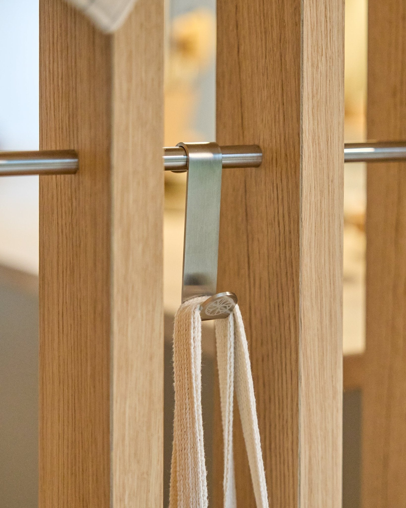 Een roestvrijstalen steel WoodUpp Alva S-Hook bevestigt een beige riem aan een stang die is gemonteerd tussen vertical houten akoestische lattenpanelen. De set bevat 2 haken.