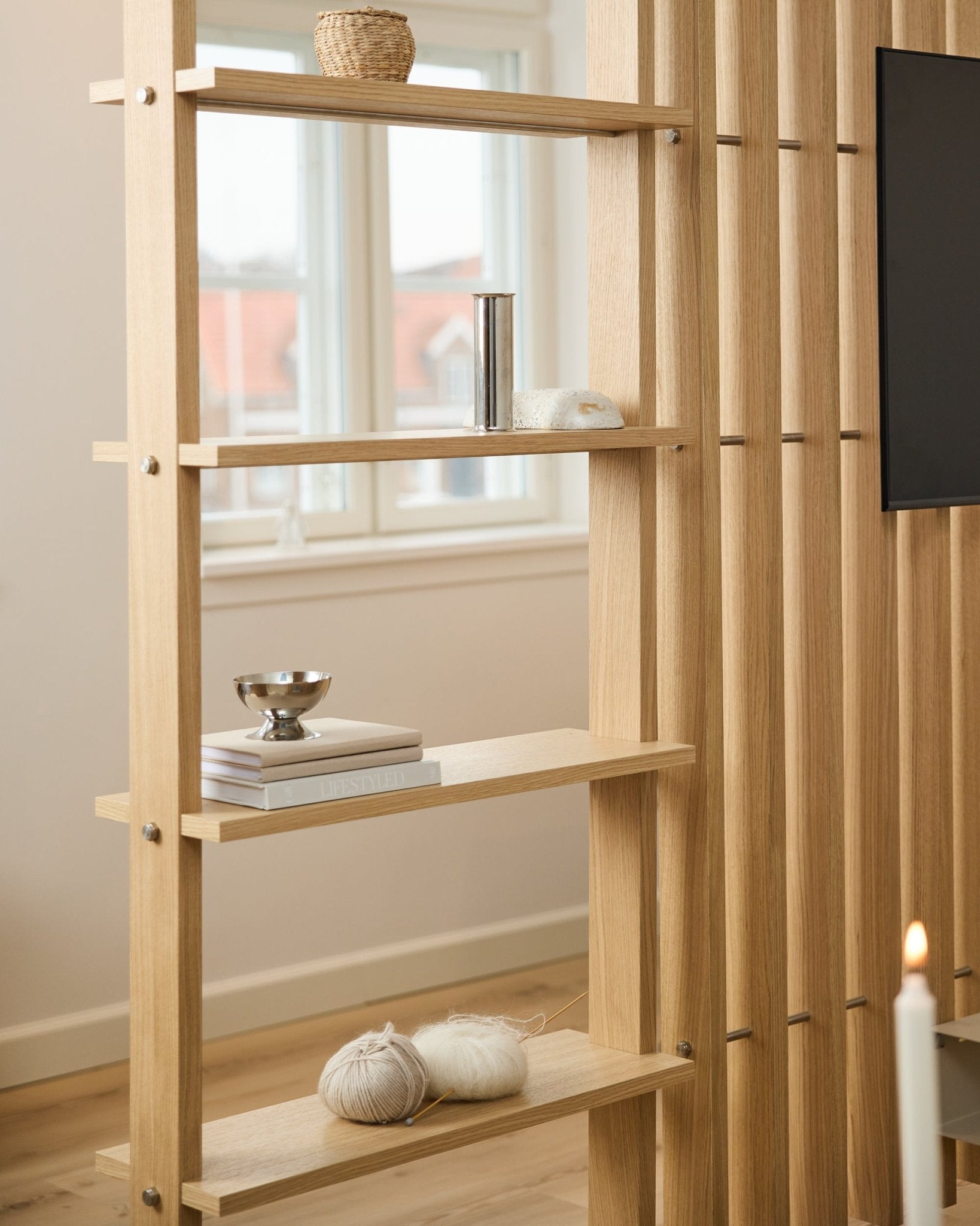 Licht eikenhouten roomdivider van WoodUpp, met vertical latten en planken met boeken en breigaren.