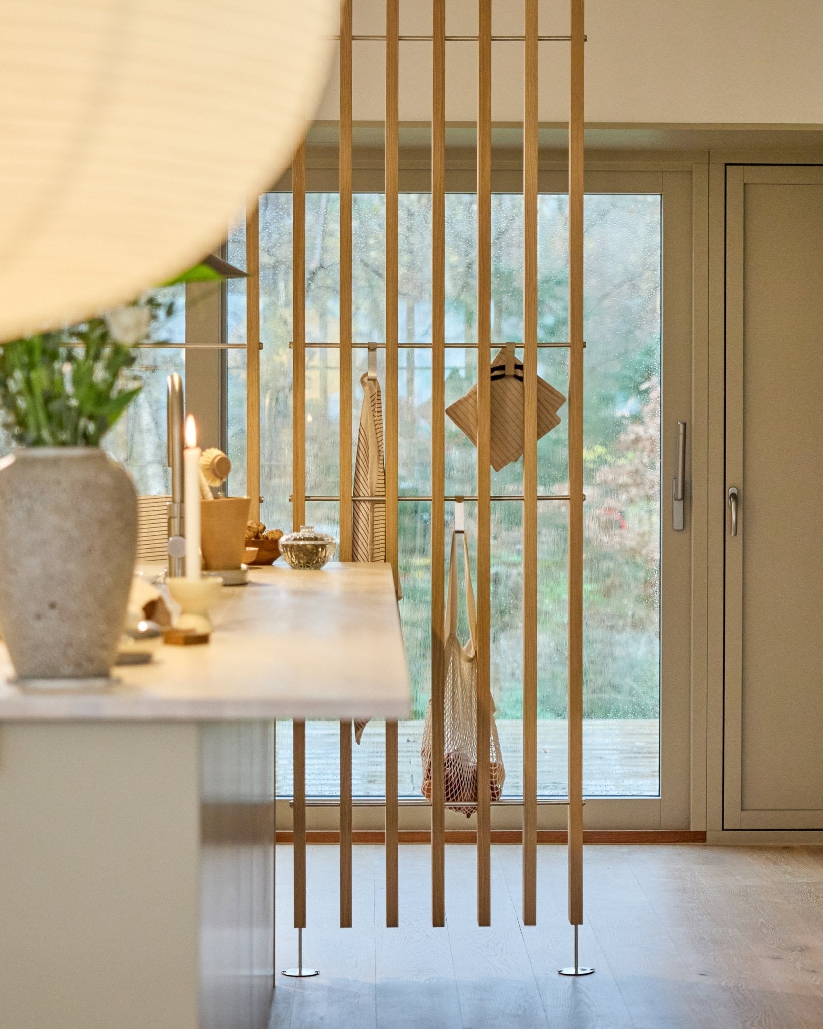 Cuisine minimaliste avec un séparateur de pièce WoodUpp Embrace (BM1, 286-302 cm) en placage chêne, avec plans de travail clairs, vase fleuri et vue sur le jardin.