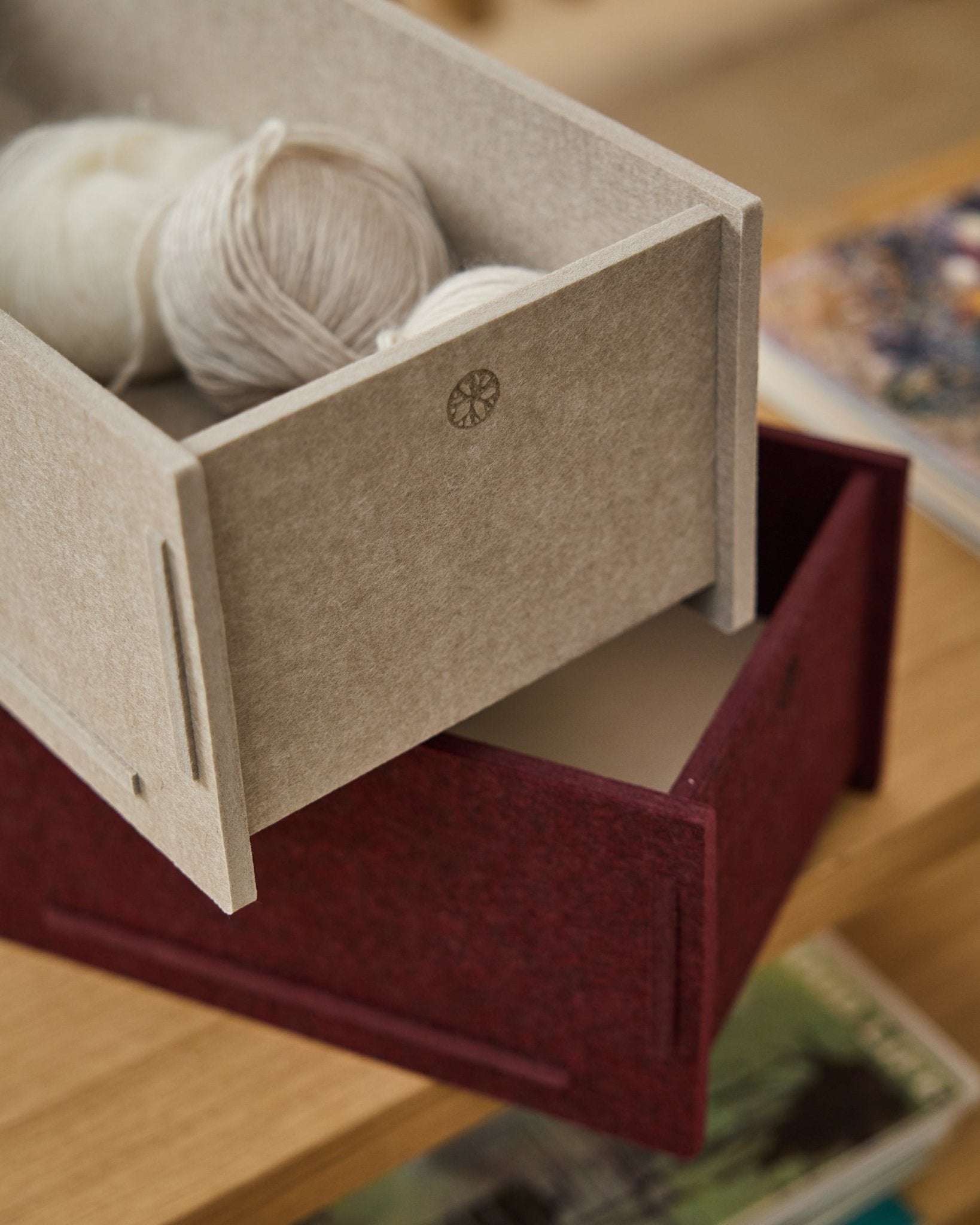 Een beige opbergbox van PET-vilt, gevuld met garen, staat boven op een gesloten WoodUpp Vigg Felt Box/BM2 in ox red, op een houten vloer met boeken op de achtergrond.