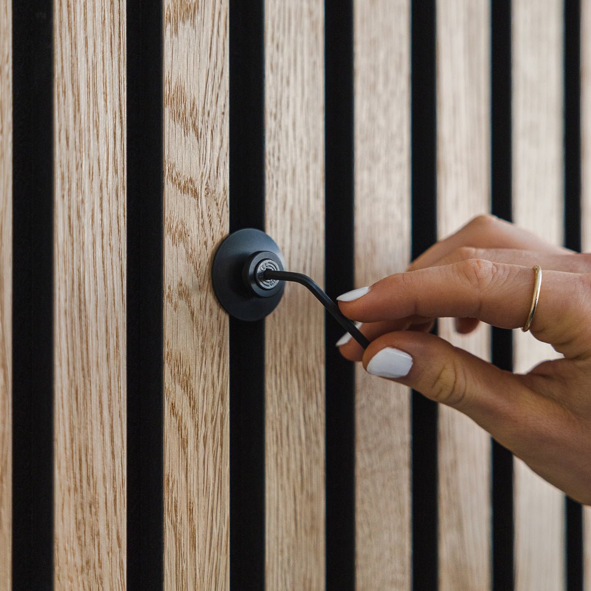 Een hand met witte nagellak draait aan een Dupps | Heavy black aluminium haak van WoodUpp op een vertical Akupanel akoestisch houten paneel.