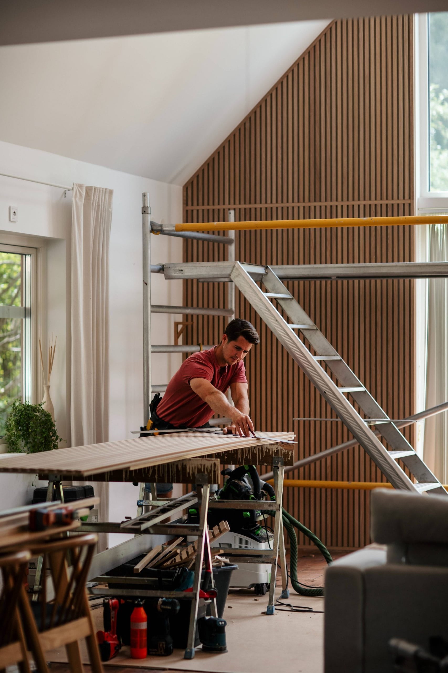 Een man meet een houten plank op een werkbank in een moderne, gedeeltelijk gerenoveerde ruimte met steigers en grote ramen, wat duidt op de installatie van akoestische panelen.