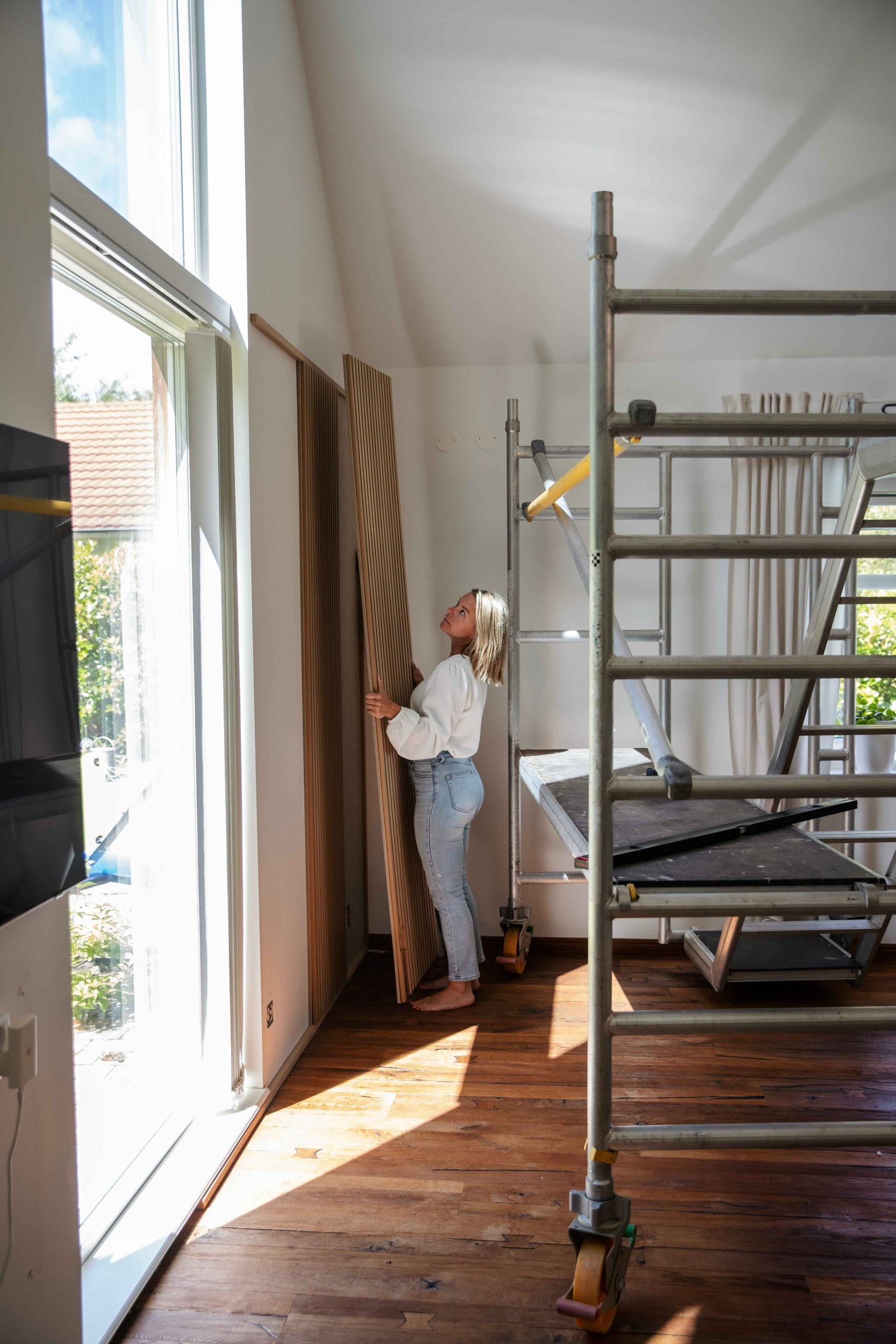 Een vrouw staat in een zonovergoten kamer met grote ramen en steigers en houdt een akoestisch houten paneel van Akupanel vast op een gepolijste houten vloer.