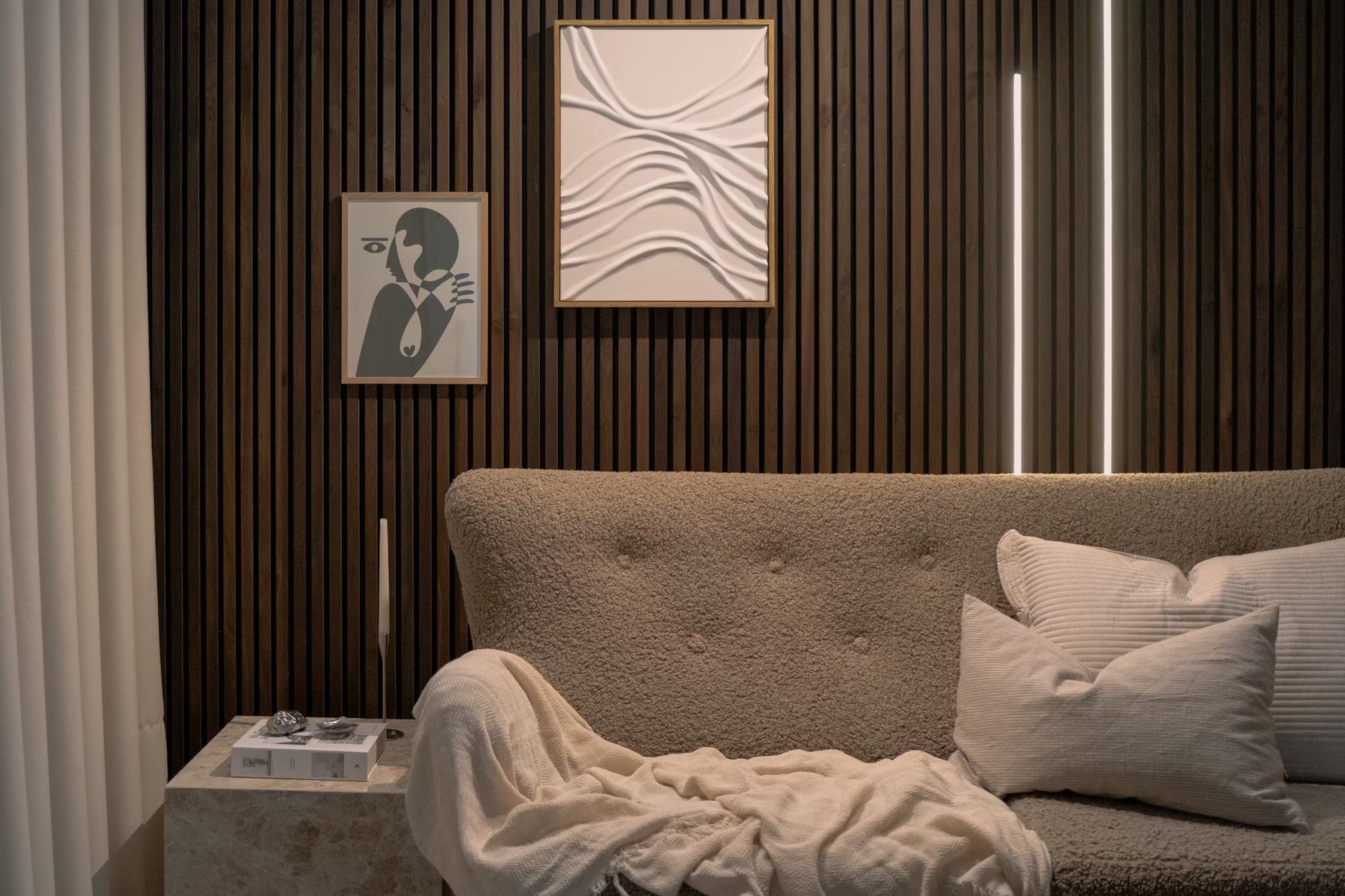 Moderne woonkamer met beige bank met textuur, witte kussens, abstracte akoestische paneelkunst en verticale Akupanel houten lattenwand met zachte sfeerverlichting.