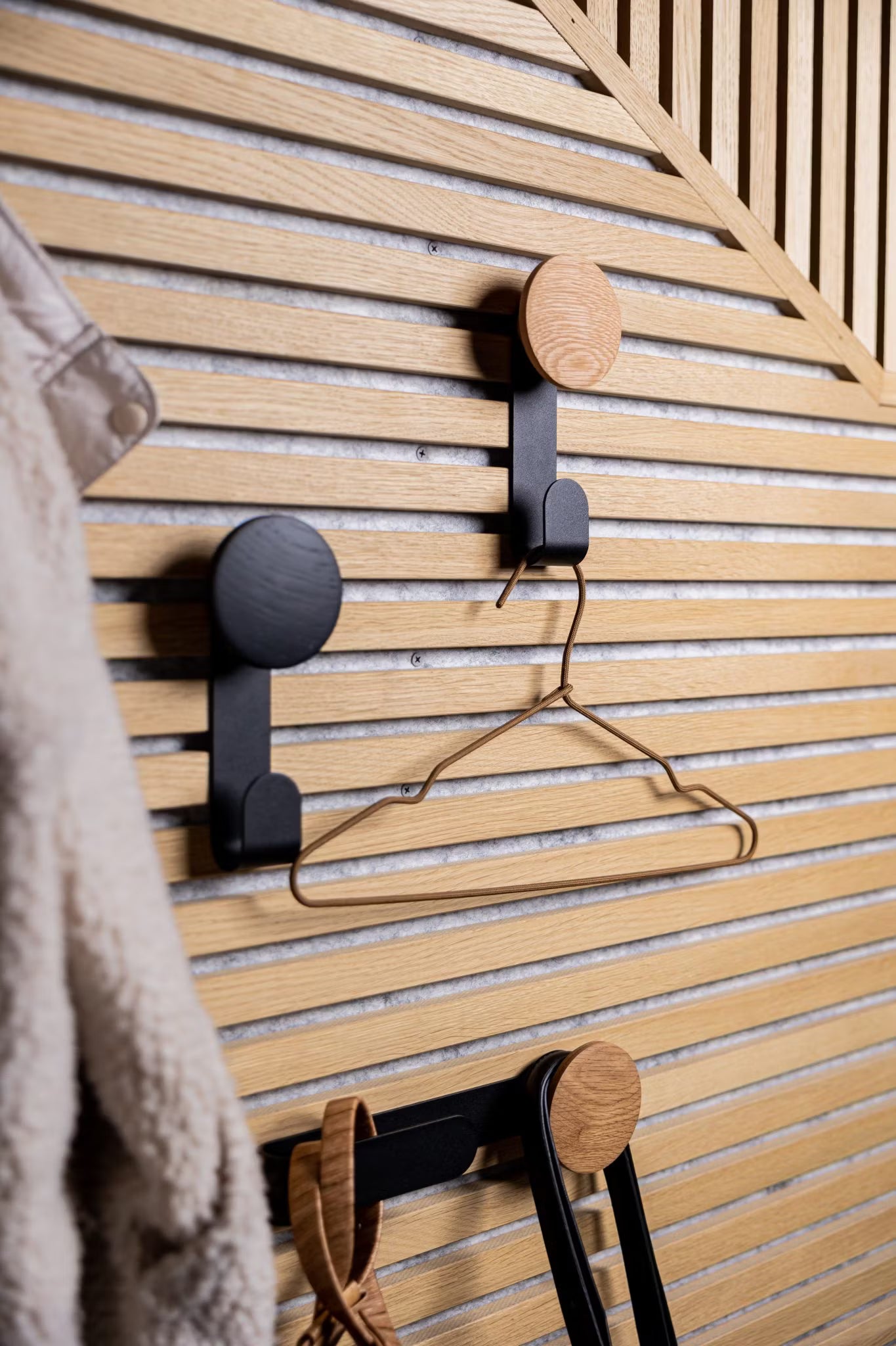 Drie moderne haken op een akoestische houten Akupanel wand in Scandinavische stijl houden een metalen hanger en accessoires vast, met links gedeeltelijk zichtbaar een pluizige jas.