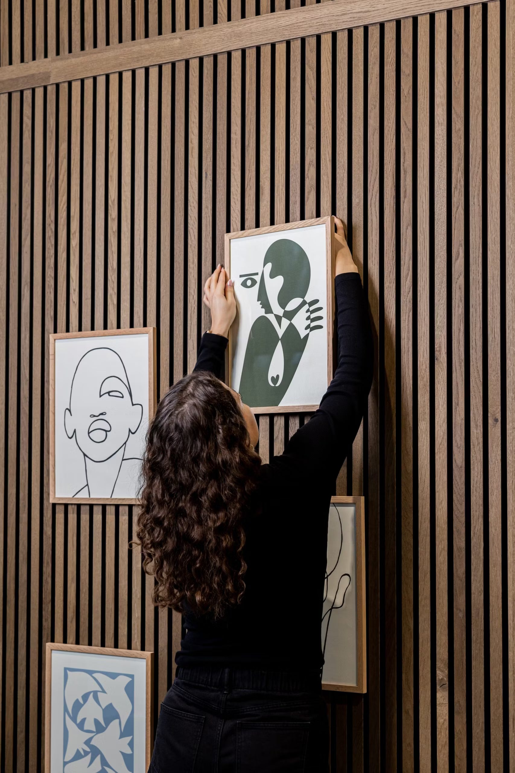 Een persoon met krullend haar hangt een WoodUpp® CREATE DUPPs accessoire aan een houten akoestisch lattenwandpaneel, omringd door minimalistische kunst en moderne decoratie.